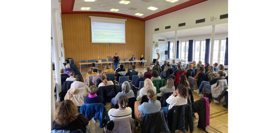 Eine Dame spricht vor mehreren Menschen in einem Saal