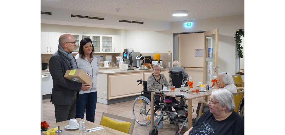 Zwei Personen stehen mit Geschenken in einem Raum - mehrere Seniorinnen und Senioren hören aufmerksam zu