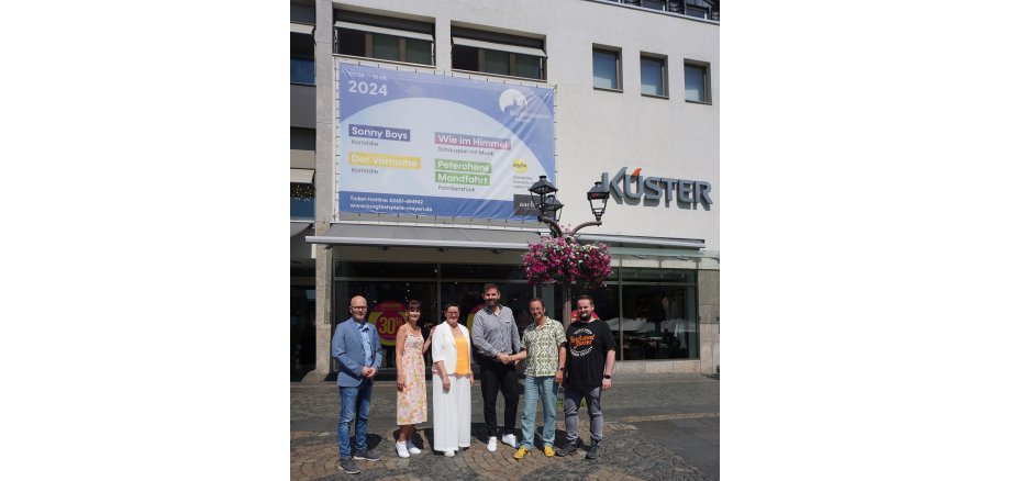 v. l. n. r.: Oberbürgermeister Dirk Meid, Leiterin der Jungen Mode Katrin Fuhrmann, Leiterin DOB Elke Münch, Leiter der Herrenabteilung Ralf Sturtz, Intendant Alexander May und Verwaltungsleiter der Burgfestspiele Kevin Stoye vor dem Plakat am Aachener Modehaus