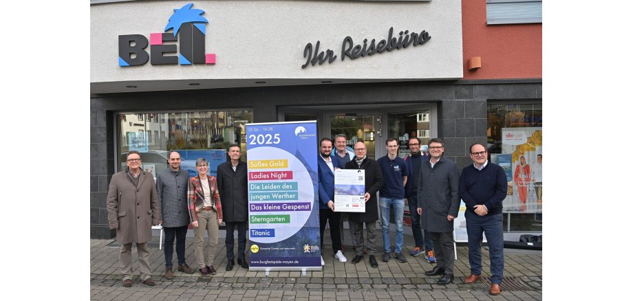 11 Personen vor dem Reisebüro Bell. Zwischen ihnen steht ein Plakat mit den Theaterstücken der Burgfestspiele 2025